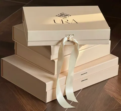 Stack of cream rigid flap-lid gift boxes with a foil logo and satin ribbon closure